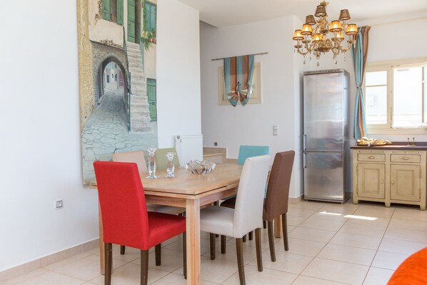 A dining area with a wooden table and chairs, a chandelier, and a kitchen with a refrigerator and cabinets. - villa victor 2 (Naxos)