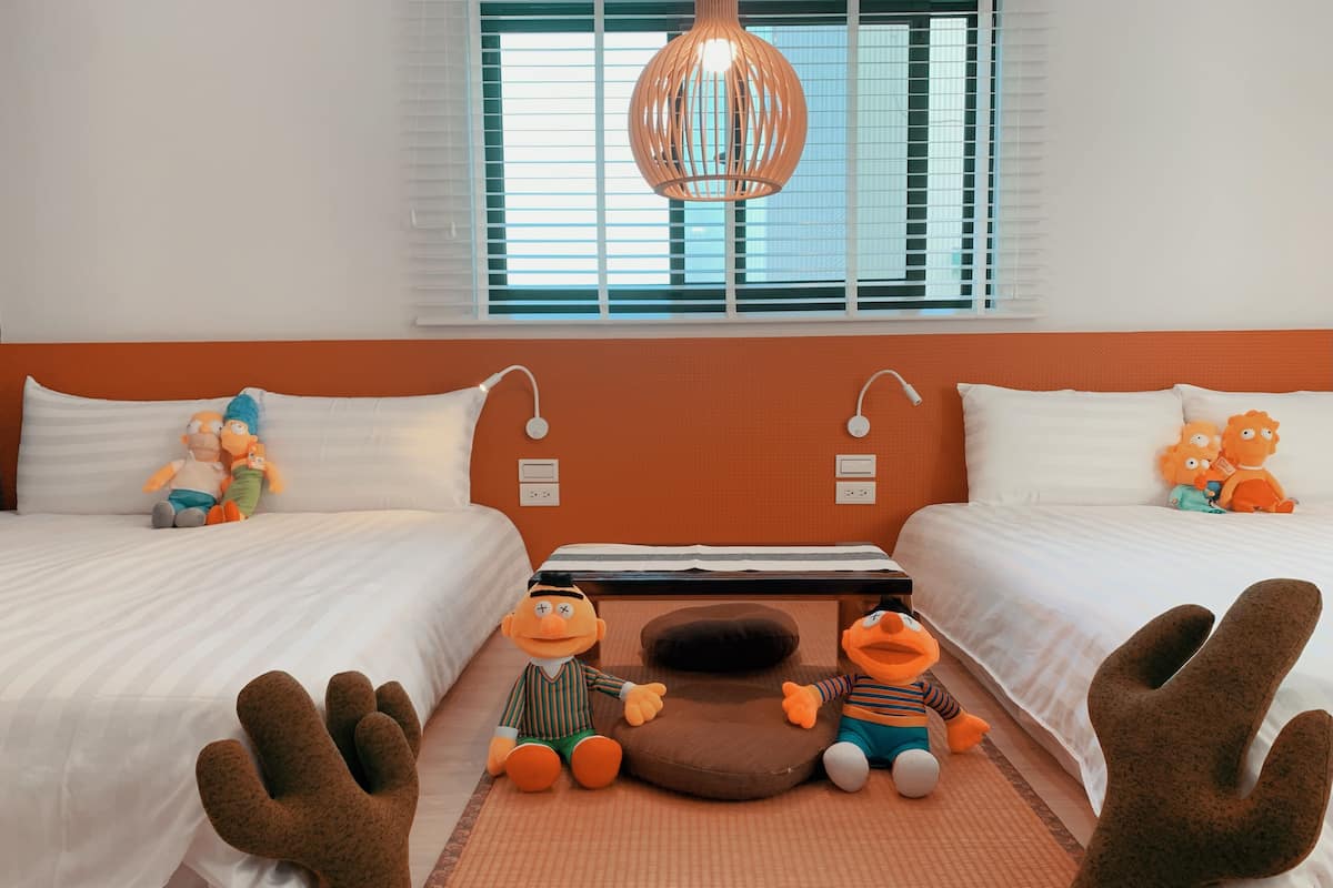 Une chambre d’hôtel avec deux lits, une grande fenêtre avec des stores, un luminaire suspendu et des peluches sur le sol.
