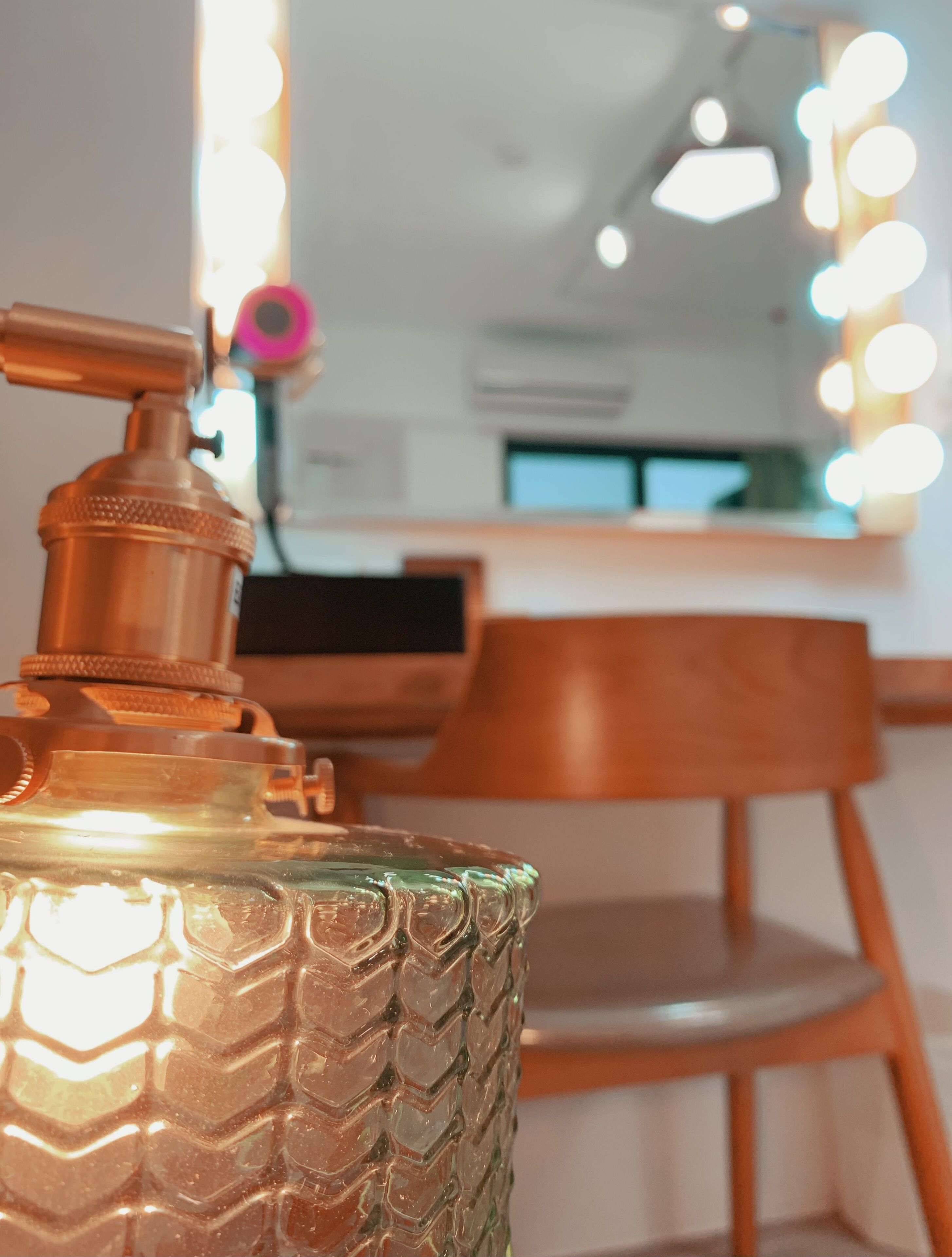 Primer plano de un recipiente texturizado de color cobre con dosificador, sobre un fondo desenfocado de un interior moderno con una silla de madera y un escritorio.