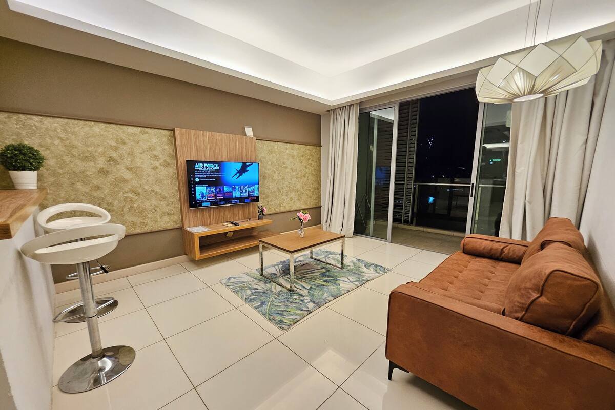 A modern living room with a flat-screen TV, a brown sofa, a coffee table, and a small bar area with stools.
