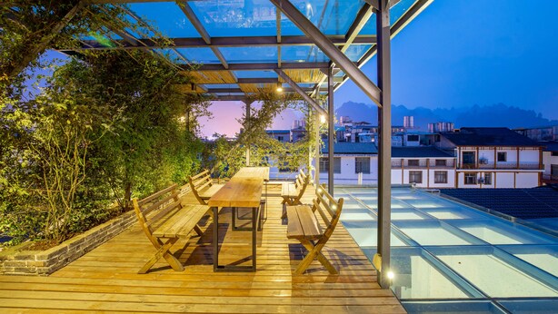 A rooftop terrace with wooden tables and benches, glass flooring, and a view of buildings in the background. - Maosao Inn (Zhangjiajie)