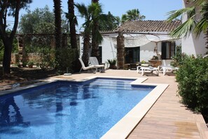Uma piscina retangular com bordas revestidas, rodeada por um deck de madeira. Há espreguiçadeiras e uma pequena mesa com toldo.