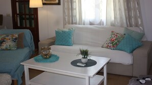 A living room with a white sofa, a small white table, and a blue armchair. There is a window with curtains, a lamp, and a framed picture on the wall. - House ideal for families with children, large garden with pool and playground (Can Picafort)