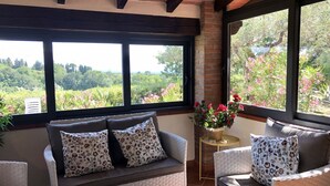 A cozy seating area with a sofa and armchairs, a small side table with a potted plant, and large windows offering a view of greenery and trees.