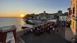 A coastal town at sunset with people dining at outdoor cafes, a historic building, and boats in the water.
