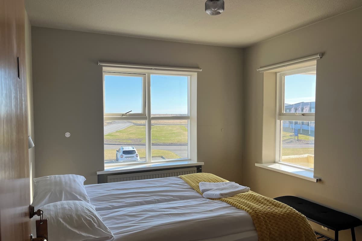 A bedroom with a bed, a bench, and two windows offering views of a road and fields.