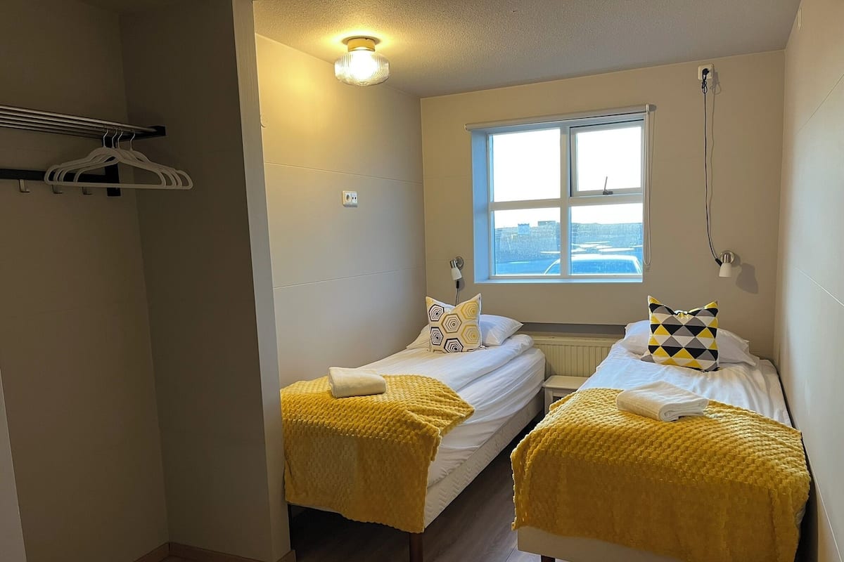 A small, bright room with two beds, a window, and a hanging coat rack.