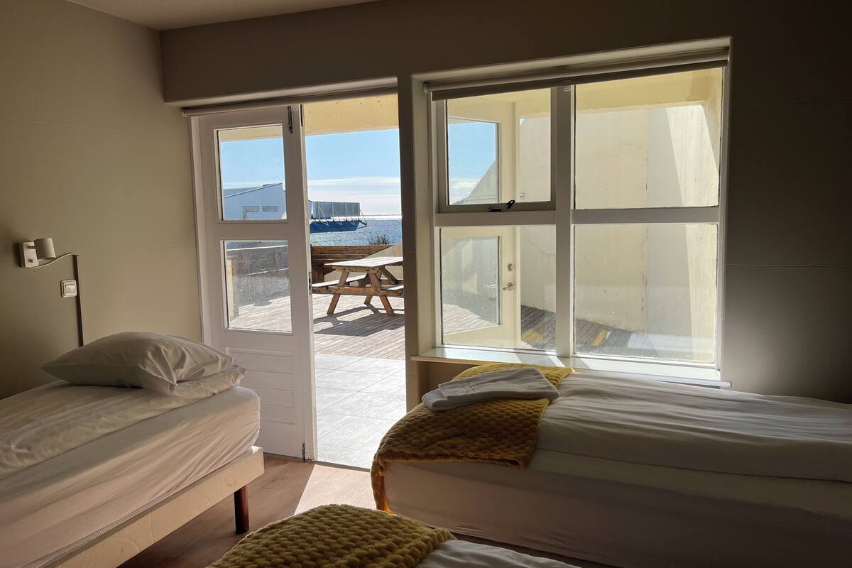A bedroom with two beds, a wooden bench, and a view of the beach through large windows.