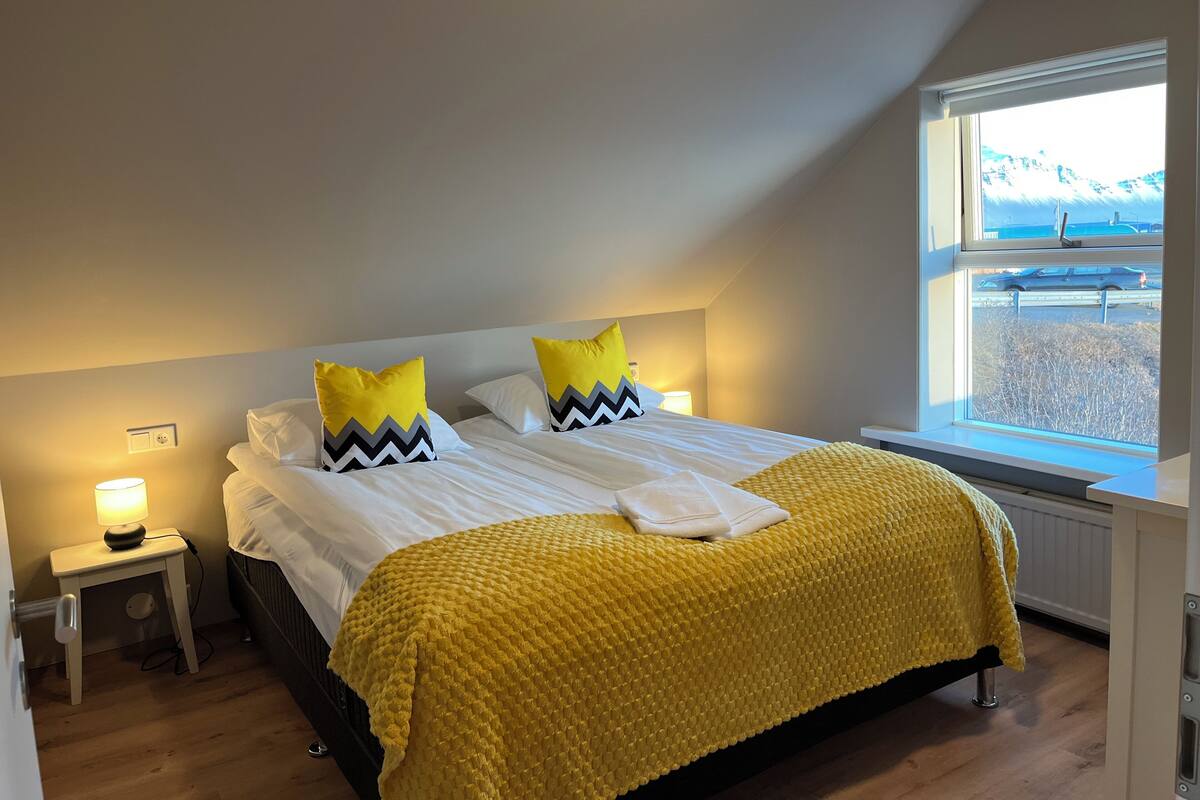 A bedroom with a bed, two pillows, a bedside table with a lamp, and a window with a view of mountains.