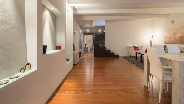 Un interior moderno con suelo de madera, una zona de comedor con mesa y sillas, una escalera y un pasillo con un espejo y elementos decorativos.