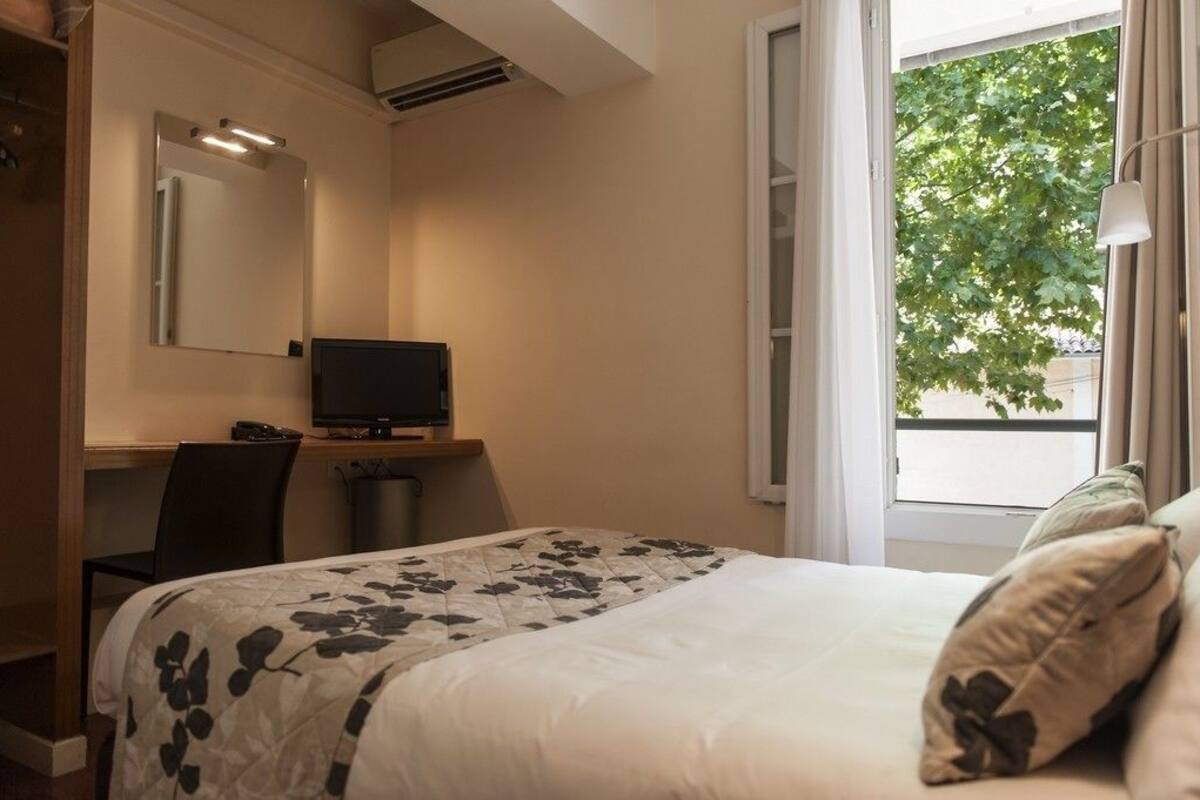 A hotel room with a bed, a desk with a computer, a chair, a TV, and a window with a view of trees.