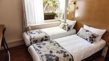 A bedroom with two beds, a window with curtains, a bedside table, and a view of greenery outside.