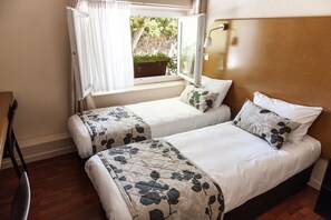 A bedroom with two beds, a window with curtains, a bedside table, and a view of greenery outside. - Hôtel Vendôme (Aix-en-Provence)