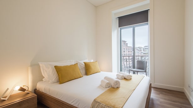 A neatly made bed with white and mustard pillows, a rolled towel, and a beige throw blanket. There is a bedside table with a lamp and a small wooden box. A window with a view of buildings is visible in the background.
