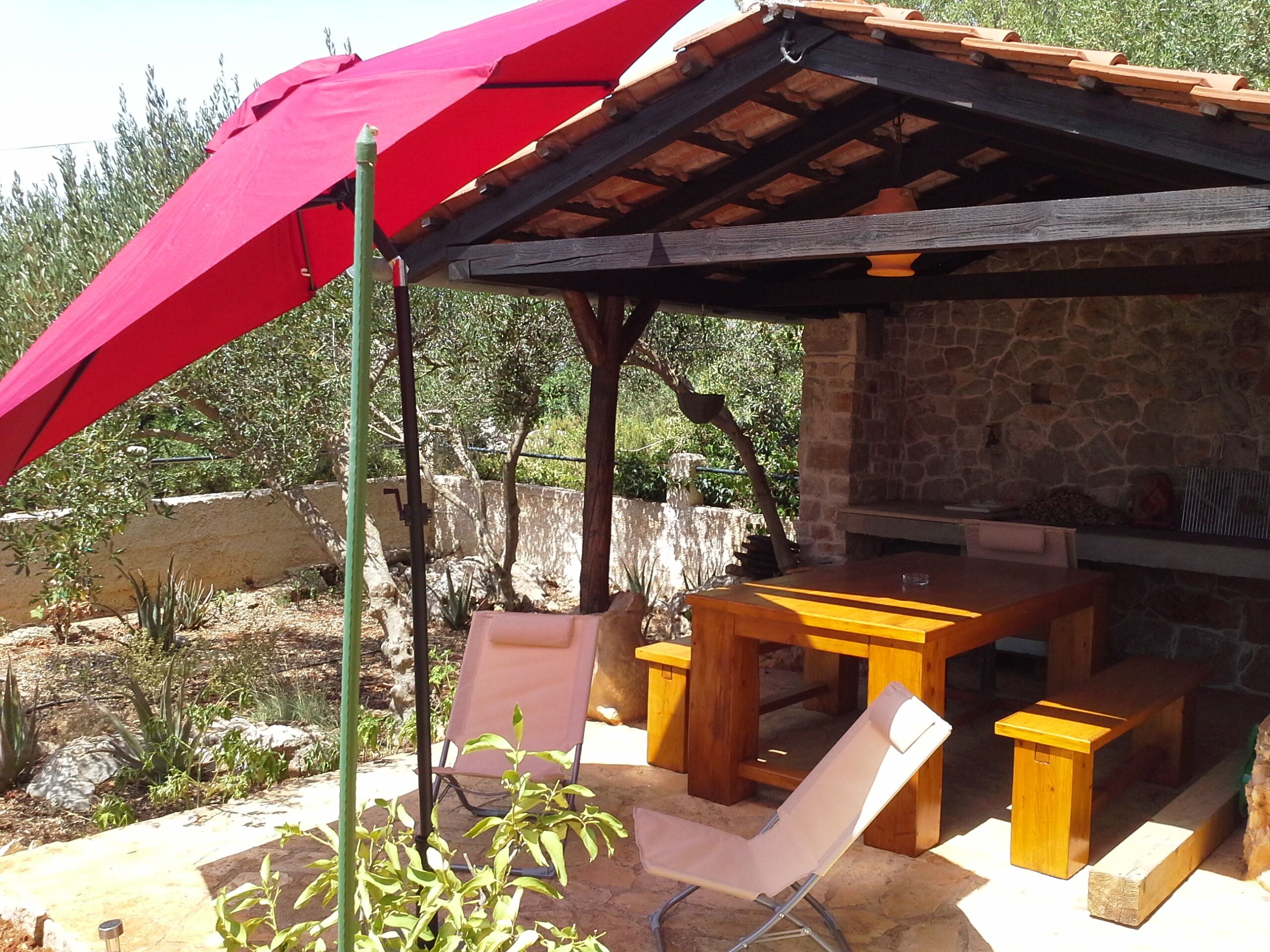 Un espace extérieur abrité, avec un mur en pierre, une table en bois et un parasol rouge.