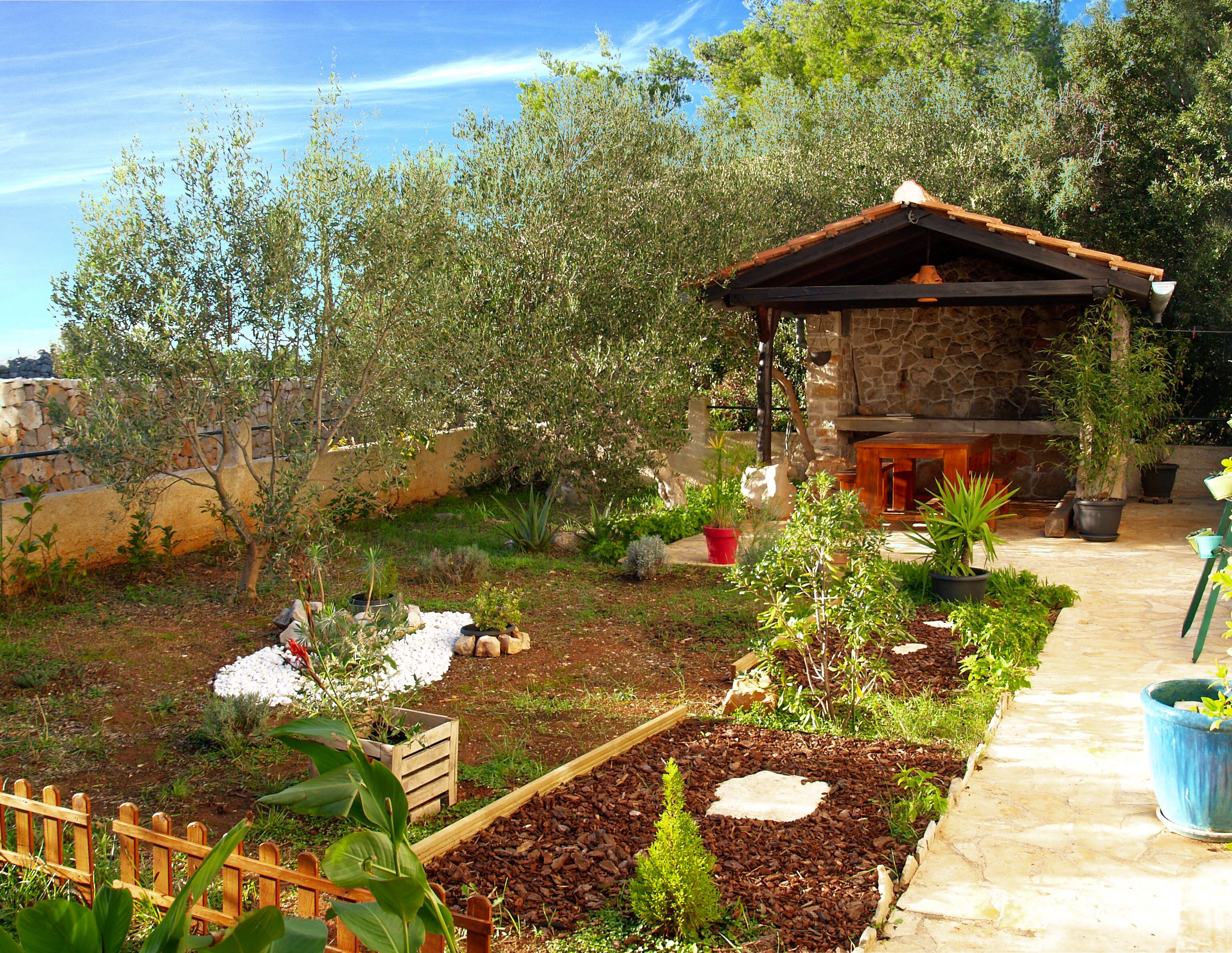 A stone building with a tiled roof, surrounded by a garden with various plants and a wooden fence.