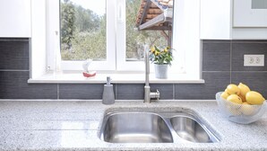 A kitchen with a double sink, a window with a view of trees, and a bowl of lemons on the counter. - Maslina Tree House (Hvar) - Peaceful retreat with garden & close to the sea (Stari Grad)