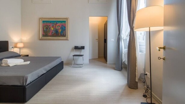 Un dormitorio con cama, lámparas de noche, una silla, un cuadro y una puerta que da a otra habitación.