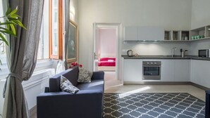 Una sala moderna con un sofá, un tapete estampado, una cocina con armarios blancos y una cama roja al fondo.