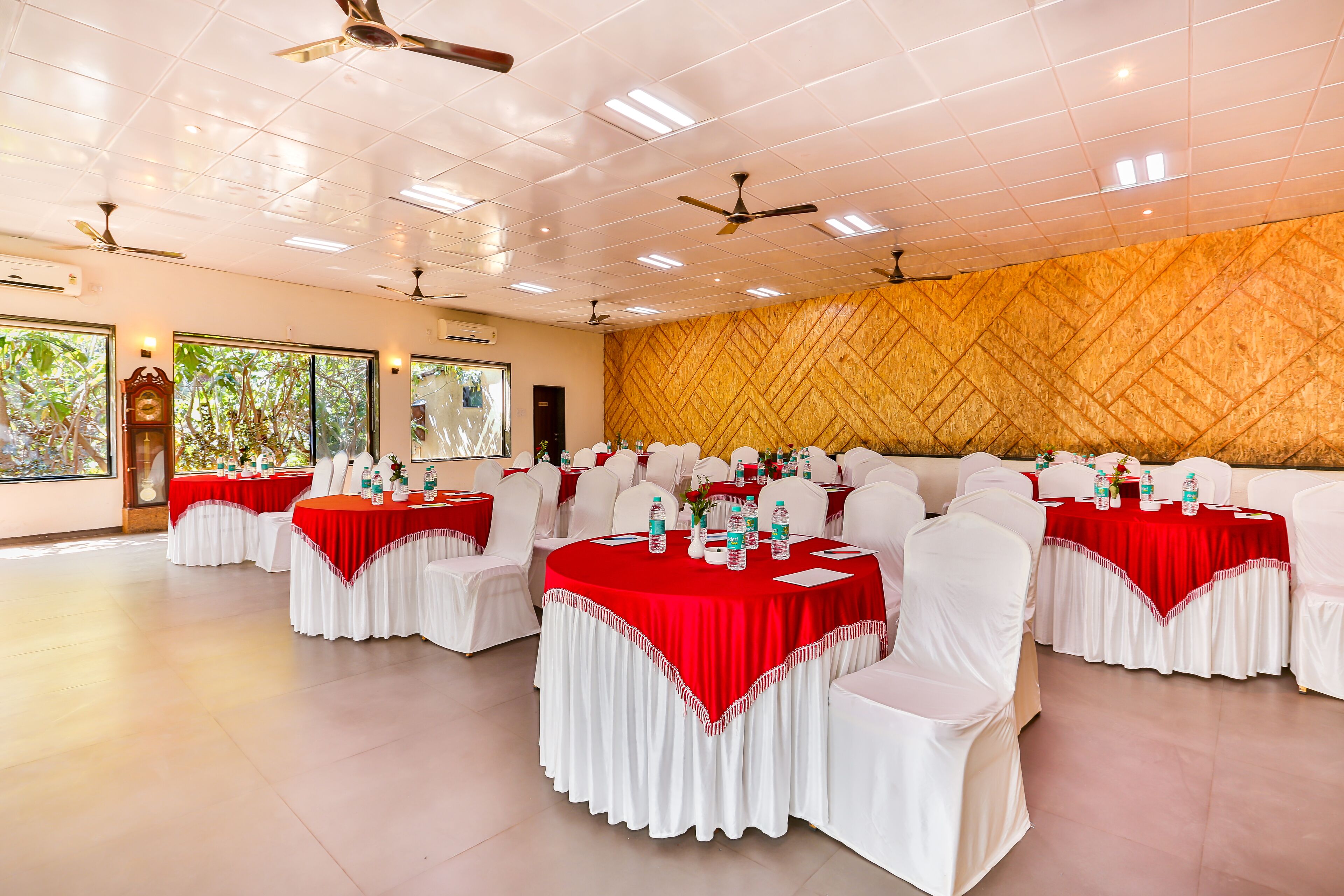 Un salón de banquetes con mesas redondas cubiertas con manteles rojo y blanco, sillas blancas, ventiladores de techo y ventanas amplias con luz natural.