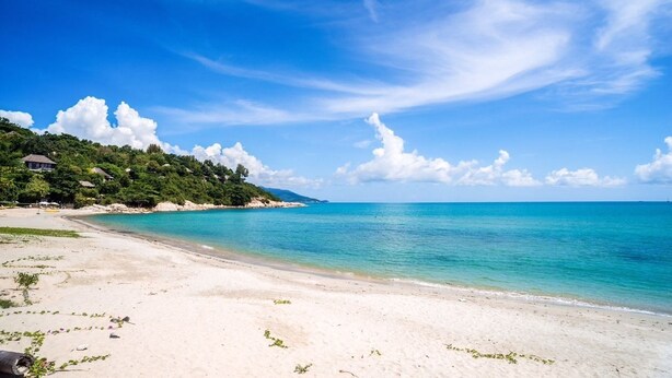 A beach with clear turquoise water, white sand, and a lush green hillside with a few buildings. - The Bay Samui (Koh Samui)