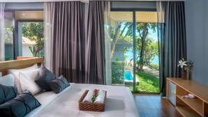 A modern hotel room with a large bed, a bedside table, a small desk, and a view of a swimming pool and greenery. - The Bay Samui (Koh Samui)