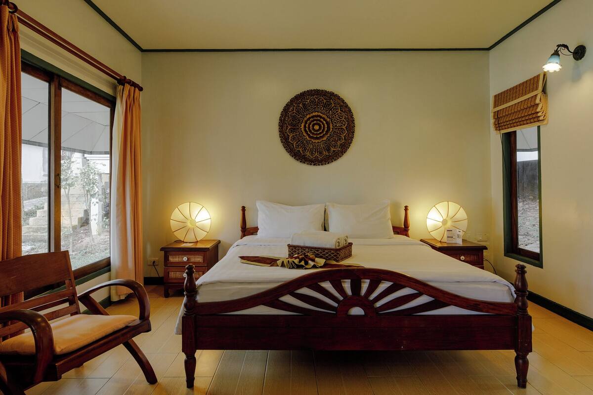 A neatly made bed with a wooden headboard, two bedside lamps, a decorative wall hanging, and a view of the outdoors through a window.