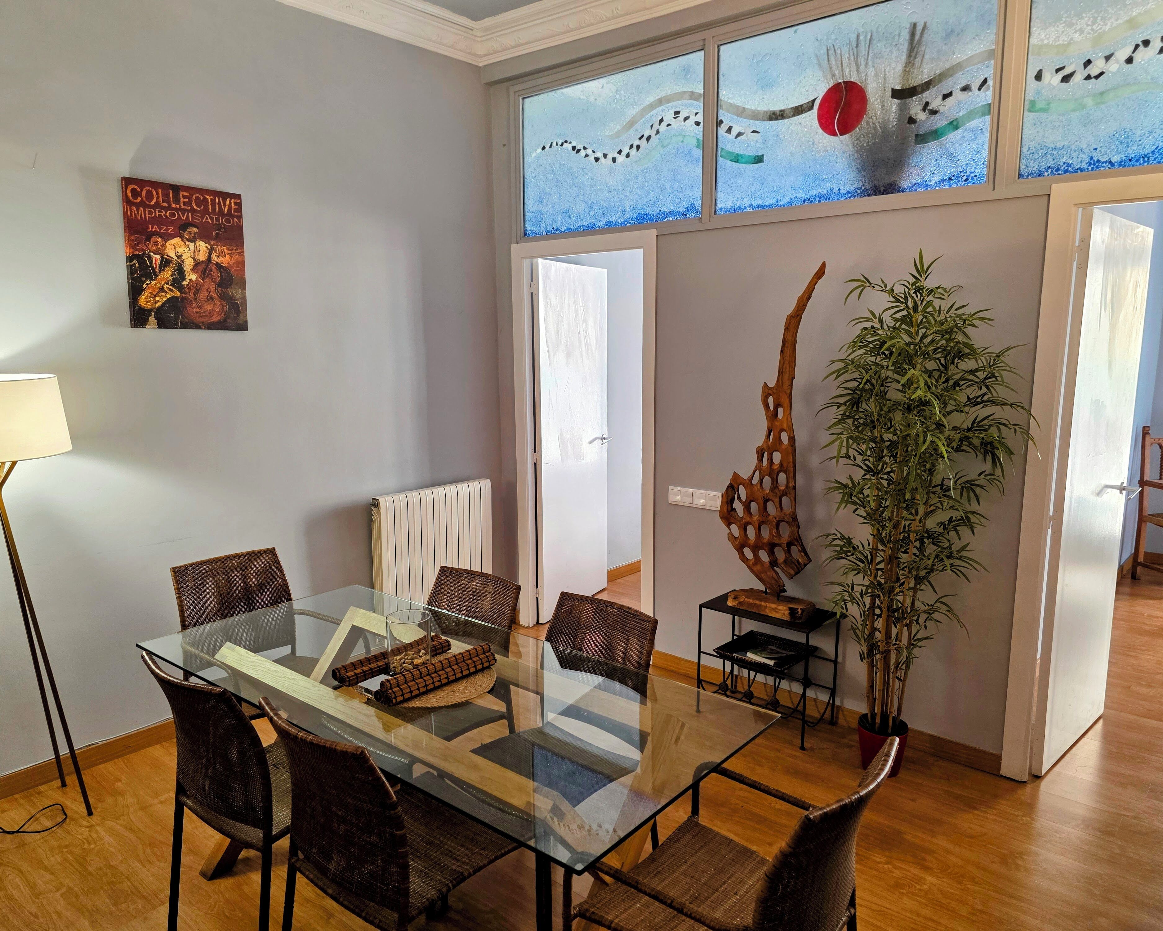 Un comedor con una mesa de vidrio y sillas de mimbre, un radiador, una escultura decorativa de madera y un póster en la pared.