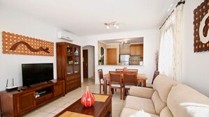 Un salon comprenant un canapé, un téléviseur, un meuble de rangement en bois et un coin repas avec des chaises et une table.