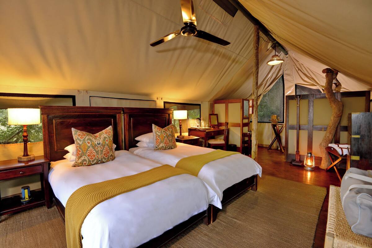 Een ruime tent met twee grote bedden, houten hoofdeinden en een plafondventilator.