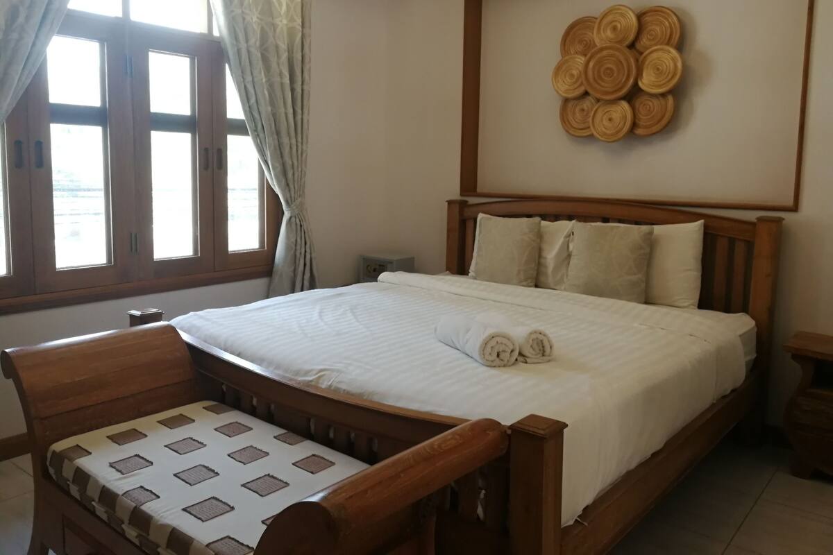 Une chambre à coucher avec un lit en bois, une table de chevet, une chaise, une fenêtre avec des rideaux et un objet décoratif fixé au mur.