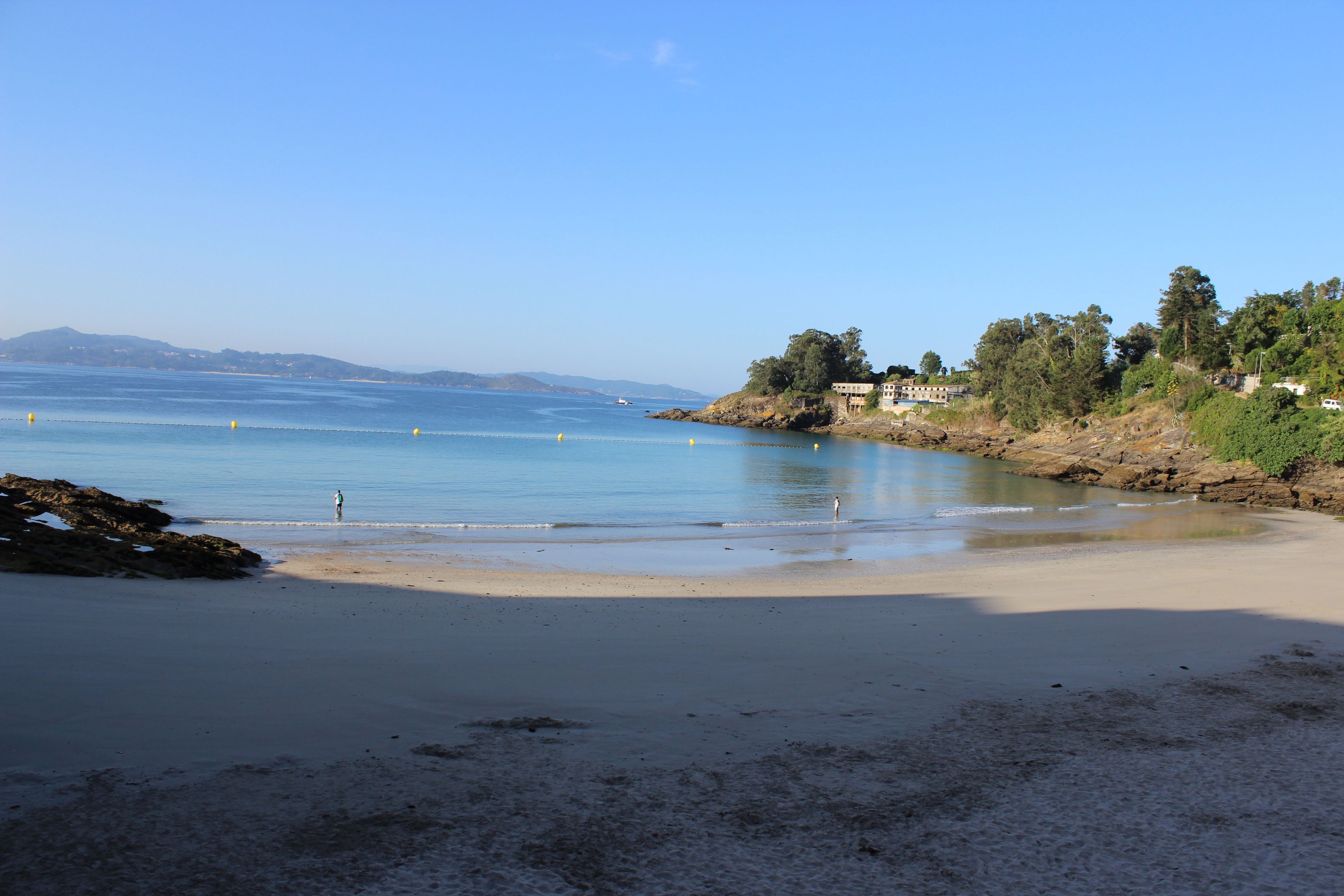 Una playa con aguas cristalinas, una orilla de arena y edificios al fondo.