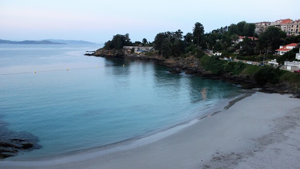 A coastal beach with a sandy shore, calm water, and buildings on a hillside. - Hotel Aquaria Caneliñas (Sanxenxo)