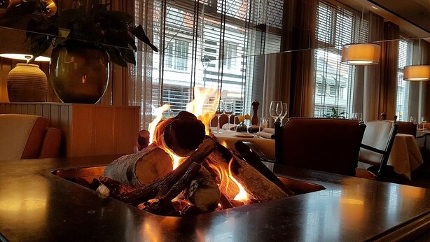 A modern dining area with a central fire feature, comfortable seating, and large windows providing a view of the cityscape. - Hotel Restaurant De Eenhoorn (Oostburg)