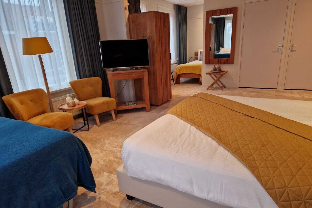 Ein Hotelzimmer mit einem großen Bett, einem Fernseher auf einem Holzständer, zwei Sesseln, einem kleinen Tisch mit einer Lampe und einem Kleiderschrank.