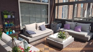 An outdoor seating area with wicker sofas and cushions, a metal shelf with potted plants, and a brick floor. - Hotel Restaurant De Eenhoorn (Oostburg)