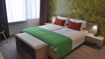 A double bed with a green bedspread, two red pillows, a wooden nightstand with a lamp, and a bench.
