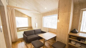 Een gezellige kamer met houten vloer, een grijze bank, een klein tafeltje en een raam met uitzicht op bomen.