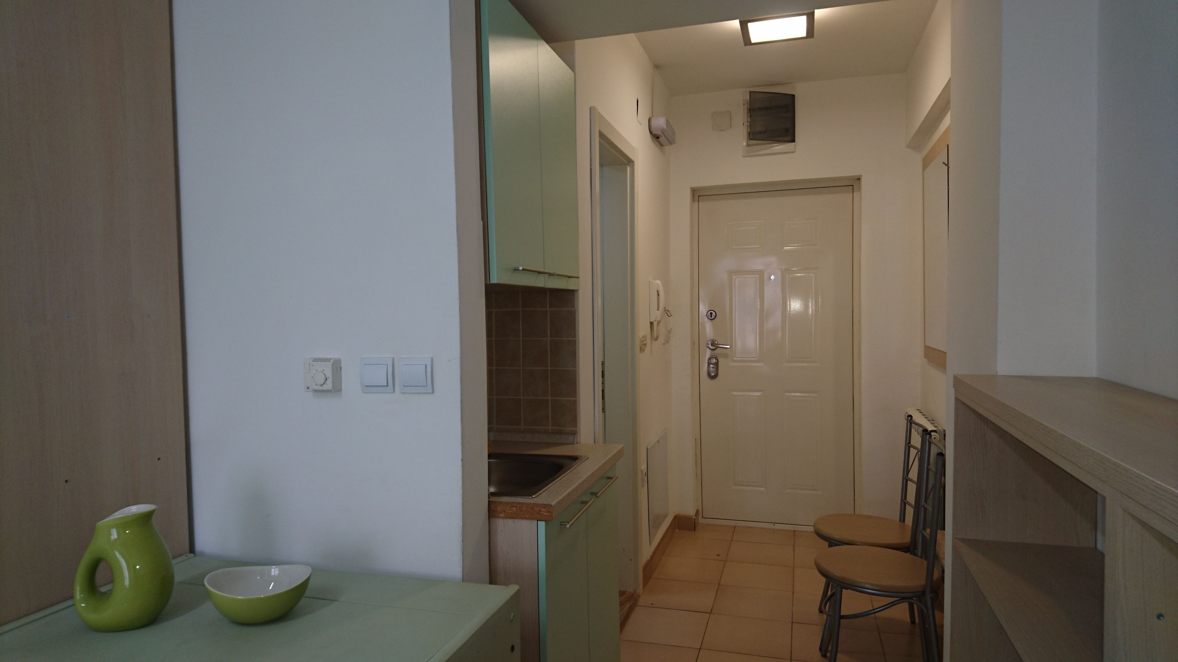 Un couloir étroit, avec un coin cuisine d’un côté, une porte au fond et un petit espace salon avec deux chaises.
