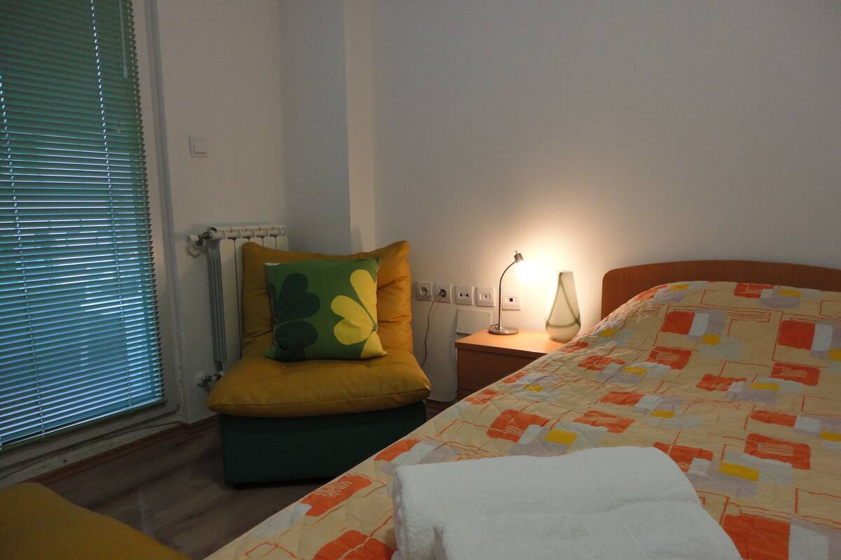 Et soveværelse med seng, et natbord, en lampe, et vindue med persienner og en stol.