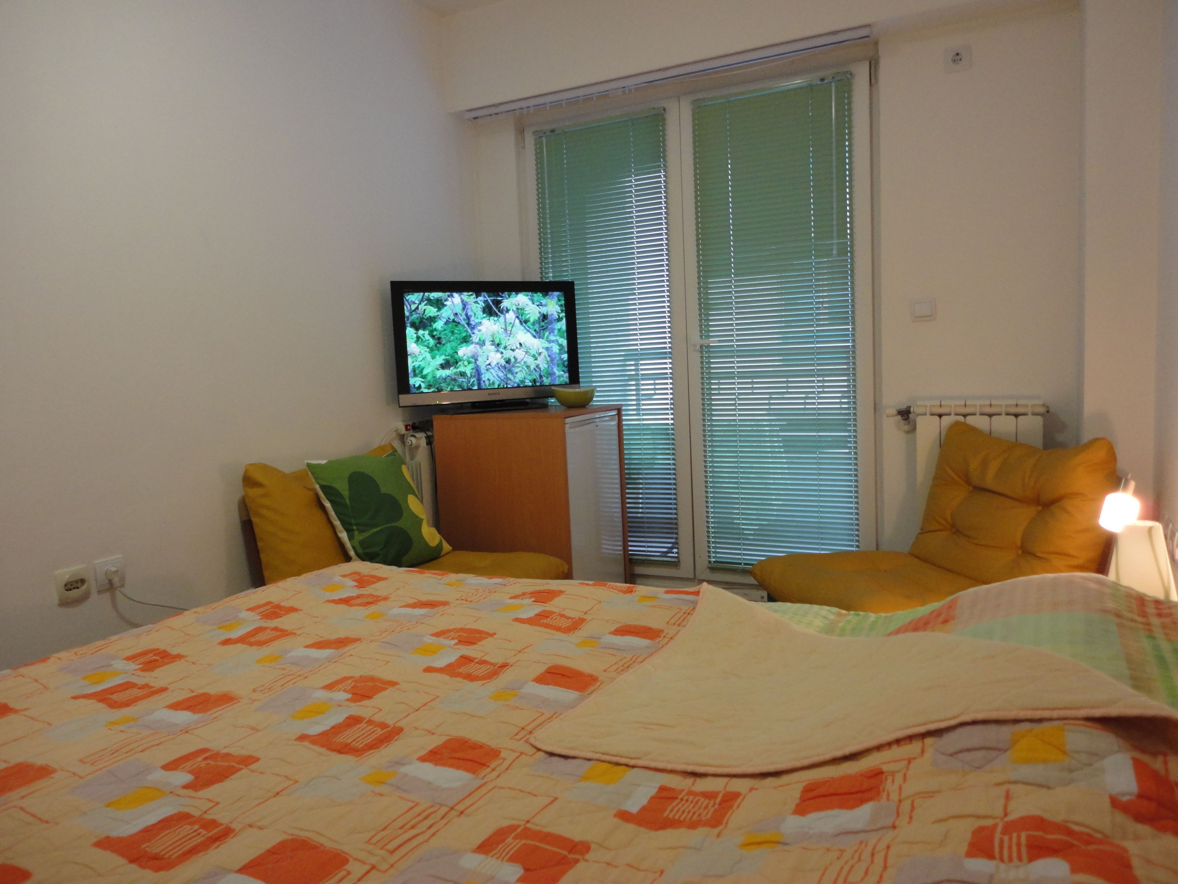 Une chambre à coucher avec un lit, un téléviseur posé sur un meuble, un radiateur et une fenêtre avec des stores.