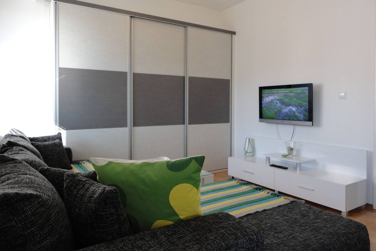 En moderne stue med en grå sofa, en pude i grøn og gul nuance, et stribet tæppe, et hvidt TV-bord og en skydelåge garderobe.