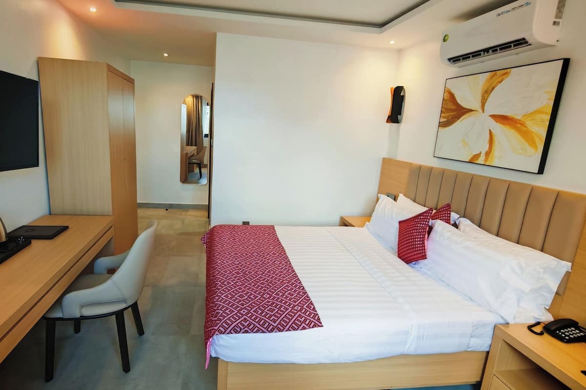 A hotel room with a bed, bedside table with a phone, a desk with a chair, a TV, a mirror, and an air conditioning unit.