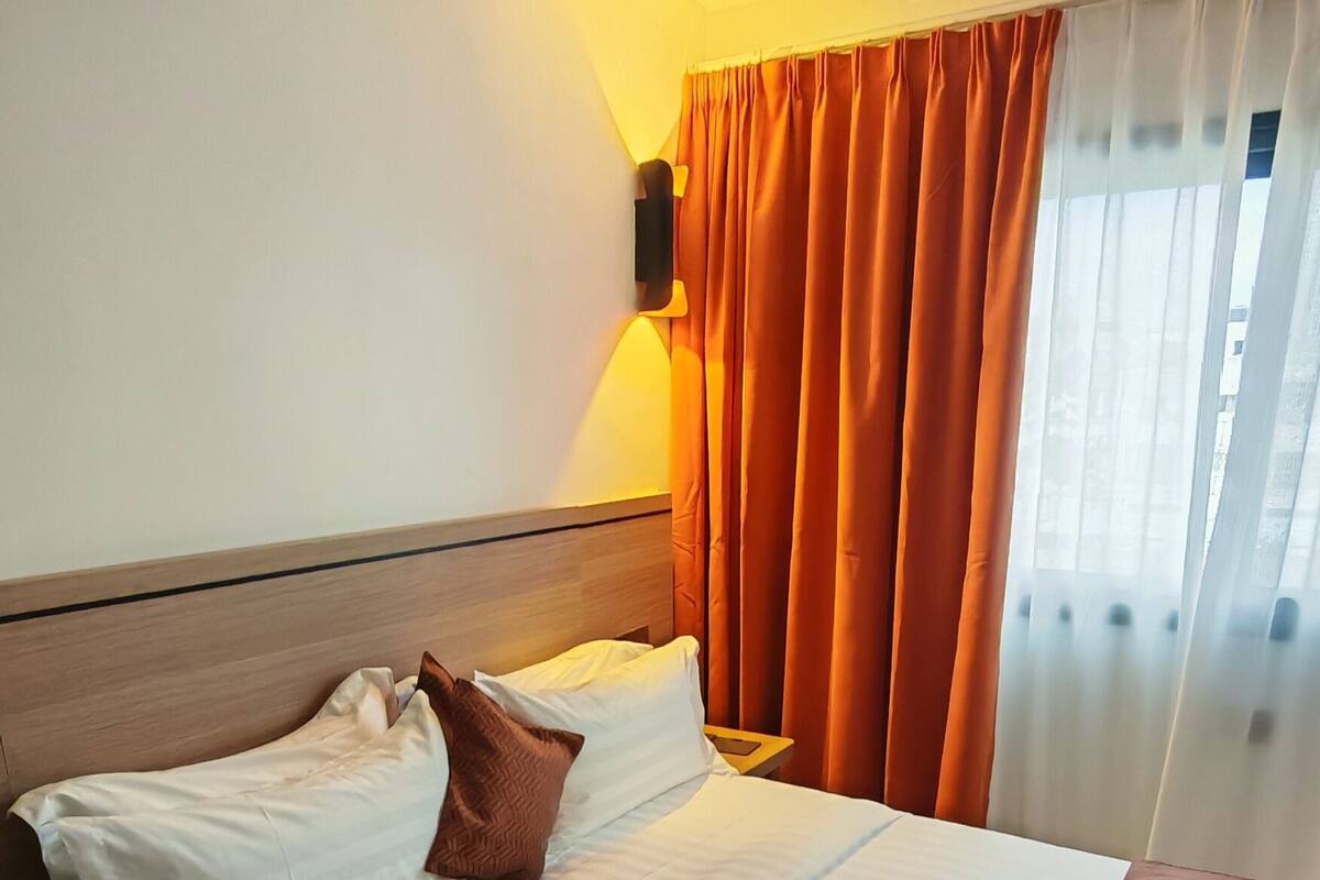 A hotel room with a bed, a nightstand, a wall-mounted air conditioner, and a window with curtains.