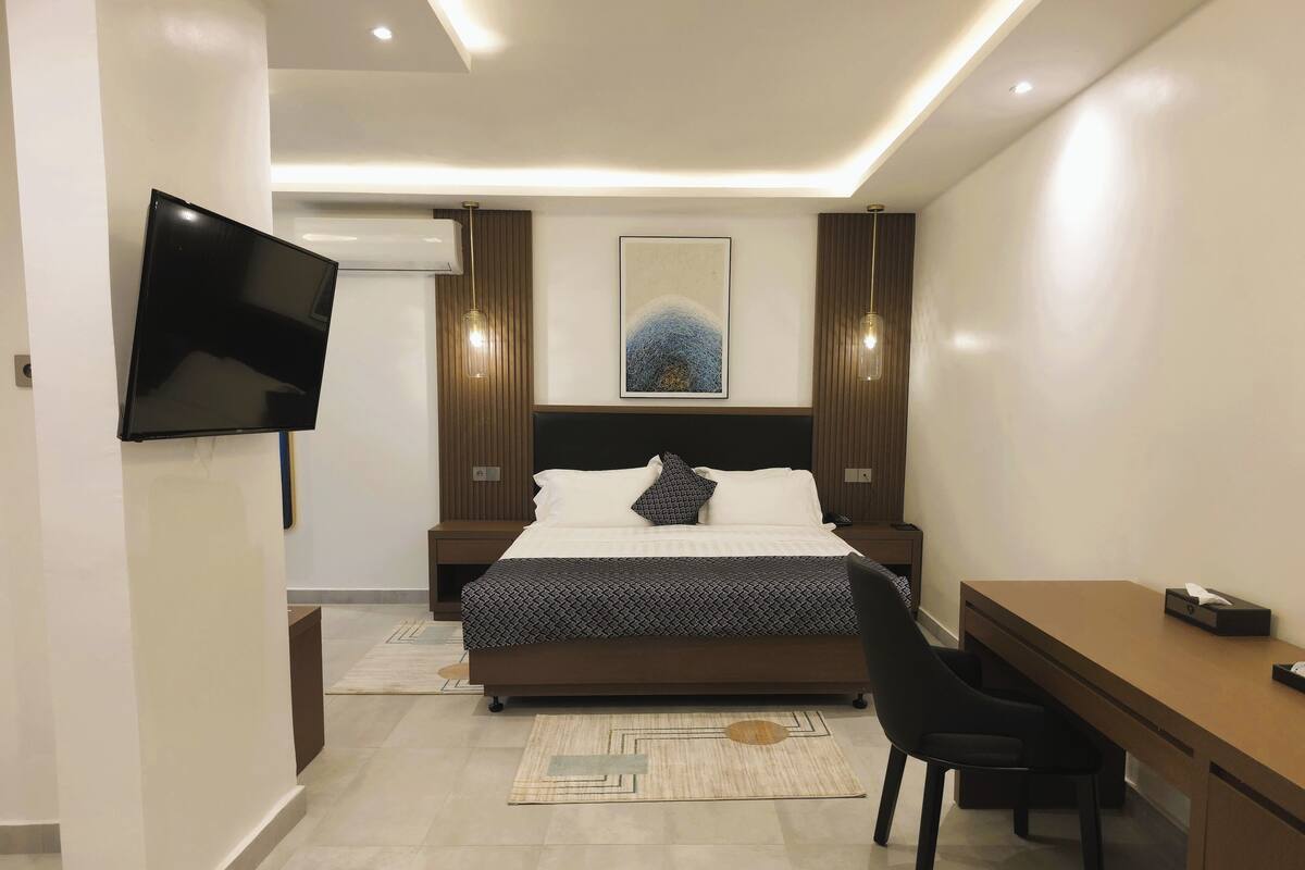 A modern hotel room with a bed, bedside tables, a desk, a chair, a flat-screen TV, and a painting on the wall.