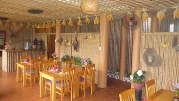 Um restaurante com mesas e cadeiras de madeira, teto de bambu e cachos de bananas pendurados.
