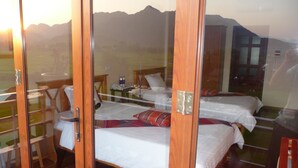 Quarto com duas camas, uma mesa de madeira e uma porta de vidro que dá para uma área externa com vista para as montanhas.