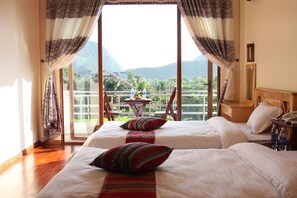 Two beds, a balcony with lush greenery views, and a small table with a vase of flowers.