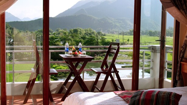 Varanda com mesa e cadeiras, uma cama e vista para uma paisagem verde com montanhas.