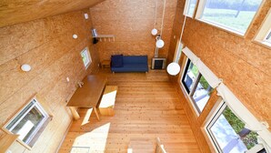 A compact, open-plan room with a wooden floor, a blue sofa, a wooden table, and large windows providing natural light. - Moon Cabin (Kutchan)